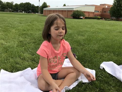 One student sits on the grass with her class as they all close their eyes in a meditation practice. 